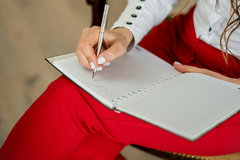 Symbolfoto: Eine Frau schreibt etwas in ein Notizbuch, das auf ihrem Schoß liegt