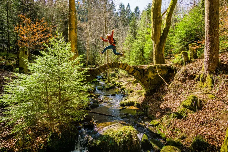 Eine Steinbrücke über einen Bach in einem Wald. Über die Brücke springt eine Frau.