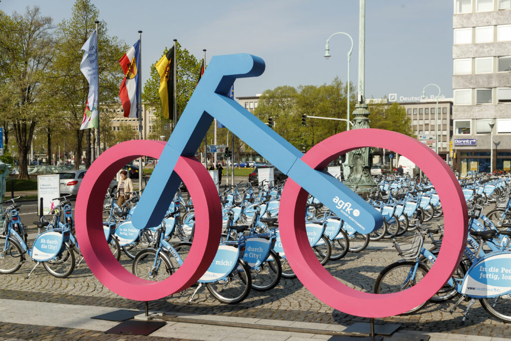 Ein riesiges stilisiertes Fahrrad in blau mit pinken Reifen steht auf einem Platz vor vielen Leifahrrädern.