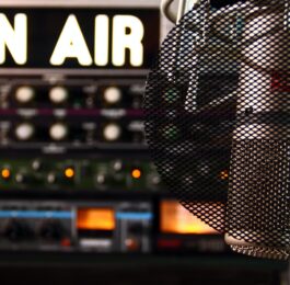 Ein Mikrofon mit Popschutz hängt von oben herab auf der rechten Bildseite. Links sieht man in großen weißen buchstaben die Worte On Air, darunter ein Mischpult.
